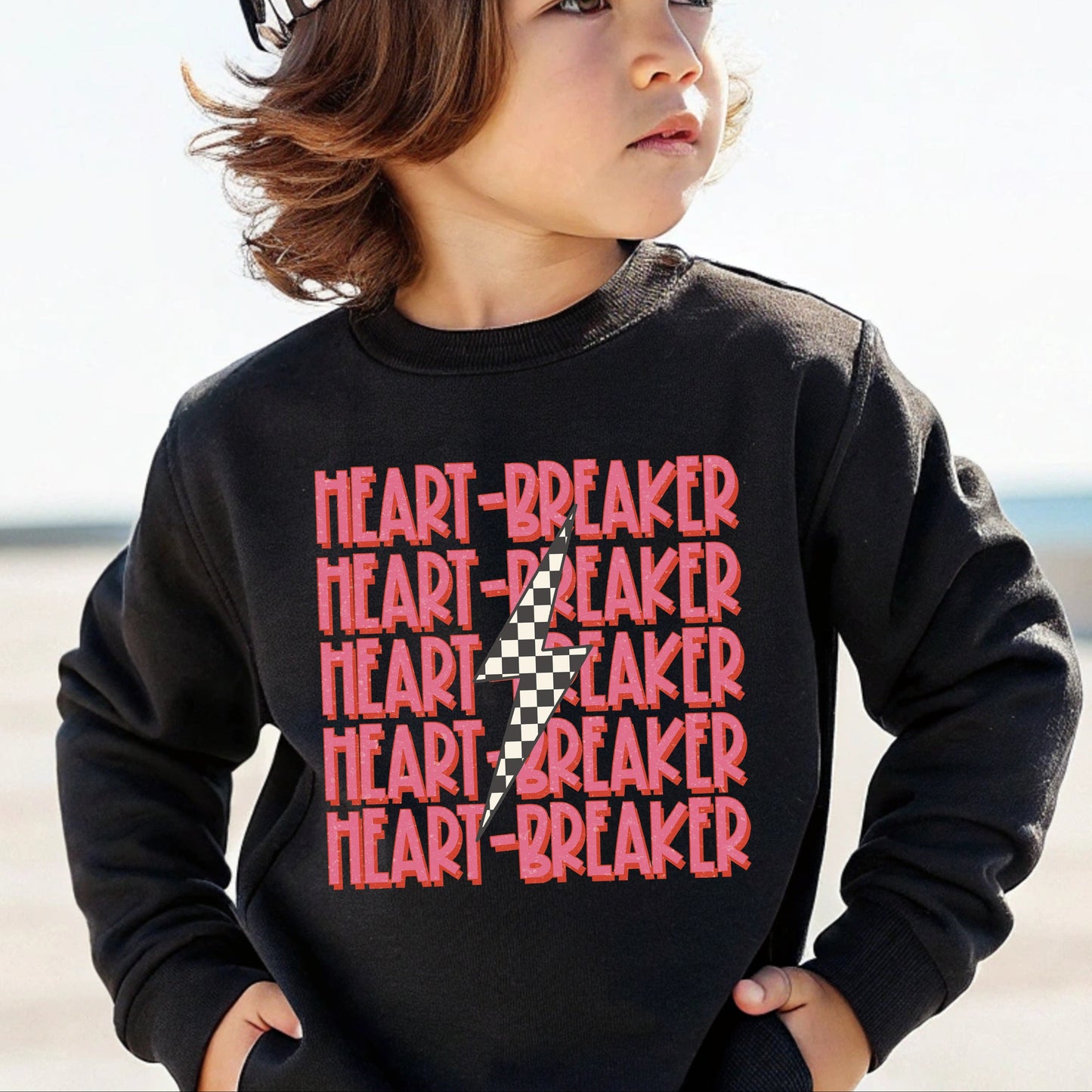 Child wearing a black sweatshirt with 'HEART-BREAKER' text and a checkered hat on a beach.