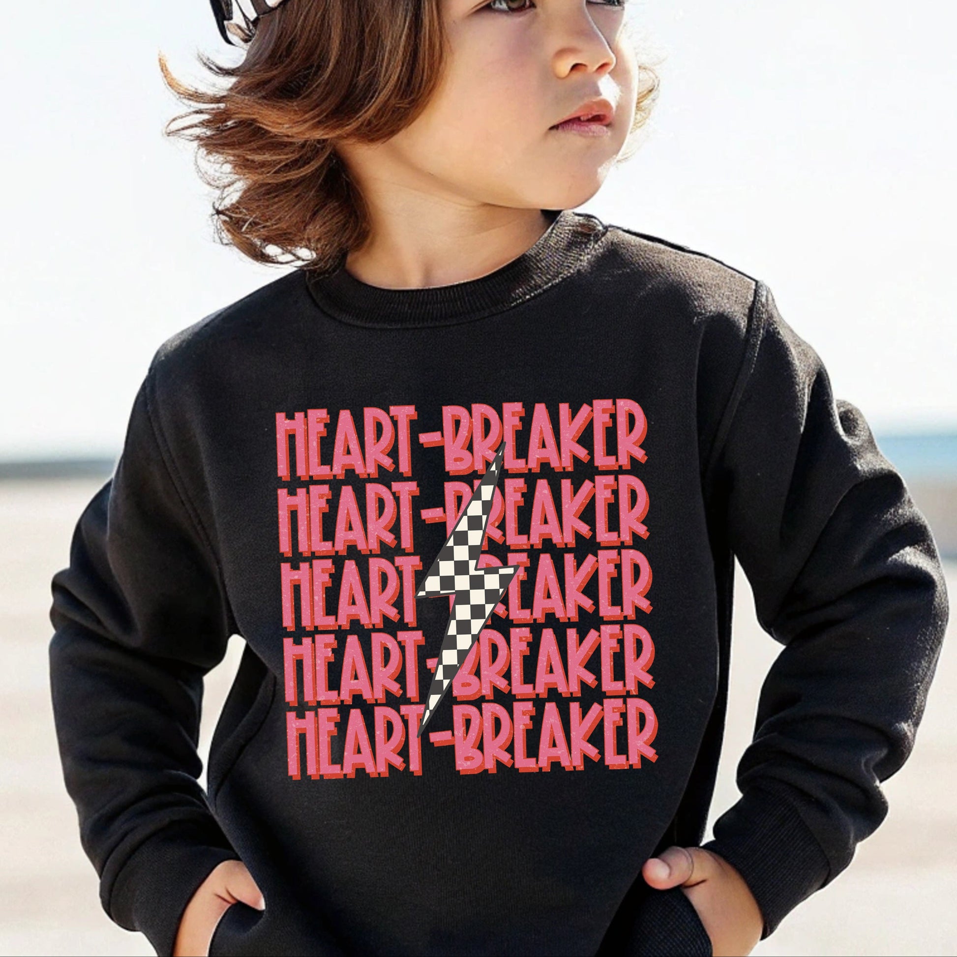 Child wearing a black sweatshirt with 'HEART-BREAKER' text and a checkered hat on a beach.