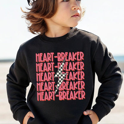 Child wearing a black sweatshirt with 'HEART-BREAKER' text and a checkered hat on a beach.