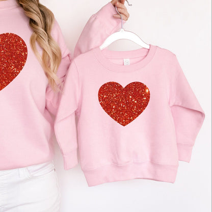 Pink sweatshirts with red glitter heart designs worn by a person and a child on a white background.